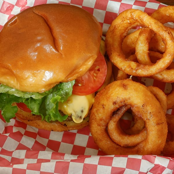 A Busta's Burger with lettuce, tomato, and cheese, served with crispy onion rings in a red checkered basket.